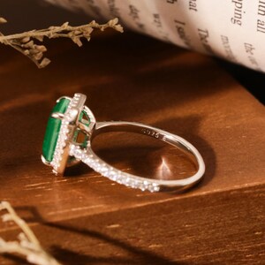 May include: A silver ring with a large emerald green gemstone surrounded by smaller clear gemstones. The ring has a band of smaller clear gemstones.