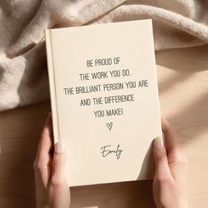 May include: A cream-colored journal held by hands, featuring the text "BE PROUD OF THE WORK YOU DO. THE BRILLIANT PERSON YOU ARE AND THE DIFFERENCE YOU MAKE!" with a heart symbol and the name "Emily". The journal is on a light-colored surface.