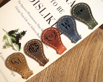 May include: A collection of leather bookmarks in various colors, including brown, red, blue, and green. Each bookmark features a unique initial or monogram design, with decorative stitching around the edges. The bookmarks are displayed on a book.