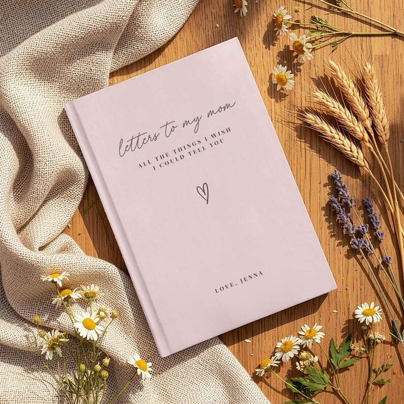 a pink book titled "Letters to my mom" with a heart symbol, placed on a wooden surface surrounded by flowers and a beige cloth.