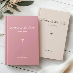 以下が含まれることがあります： ピンクとベージュの2冊のハードカバーノート。エレガントな筆記体で「Letters to the Bride」と書かれています。各ノートにはハートのシンボルと「2025年7月28日」の日付があります。結婚式用にデザインされたノートです。