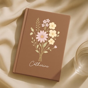 May include: A brown journal featuring a floral bouquet design. The bouquet includes pink, yellow, and white flowers with green stems. The name "Catherine" is written in cursive below the flowers.
