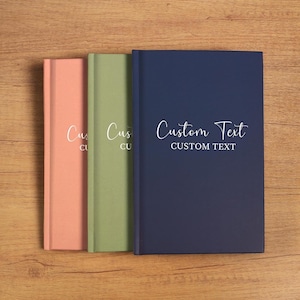 Op de afbeelding: Drie hardcover journals in koraal, olijfgroen en marineblauw. Het marineblauwe journal heeft de tekst "Custom Text" in een wit scriptlettertype. De andere twee journals hebben het woord "Custom" in een wit scriptlettertype.