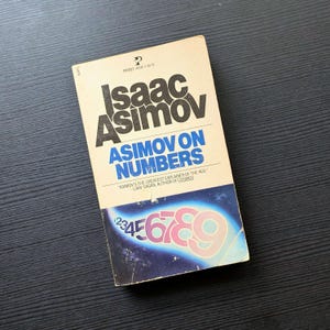 May include: A vintage paperback book titled "Asimov on Numbers" by Isaac Asimov. The cover features the author's name in large, bold letters, with the title in blue. The bottom of the cover displays numbers in pink and blue.