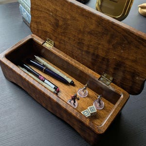 May include: A wooden box with a hinged lid, stained a warm brown. Inside, various pens and small figurines are arranged. The box is open, revealing its contents, including a few dice. The box is designed for storage and organization.