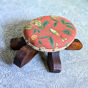 May include: A small, wooden footstool with a round, padded top. The top is covered in a red fabric with a floral and bird pattern in yellow, green, and white. The legs are dark brown and angled.