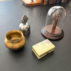 May include: A collection of objects including a small brass bowl with etched patterns, a silver-toned bell with a bird-shaped handle, a glass cloche on a dark wooden base, and a rectangular cream-colored ceramic box with a postage stamp design.