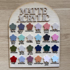 K&ouml;nnte beinhalten: Eine Holztafel mit der Aufschrift "MATTE ACRYLIC". Darunter ein Raster mit bl&uuml;tenf&ouml;rmigen Farbmustern in Farbt&ouml;nen wie Farn, Dusty Lilac und Sandstein. Die Farbbezeichnungen sind darunter aufgedruckt. Metrische Ma&szlig;e.