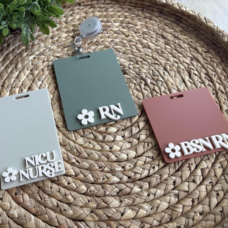 K&ouml;nnte beinhalten: Drei rechteckige Ausweishalter in Grau, Gr&uuml;n und Mauve, jeweils mit wei&szlig;en floralen Akzenten und Text. Das gr&uuml;ne Abzeichen lautet "RN", das mauvefarbene Abzeichen lautet "BSN RN" und das graue Abzeichen lautet "NICU NURSE". Eine einziehbare Ausweisrolle ist angebracht.