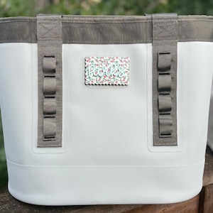 May include: A white tote bag with grey trim and two vertical grey webbing straps featuring multiple loops. A rectangular patch with a scalloped edge and a floral pattern displays the word parks in light green script lettering on the front.