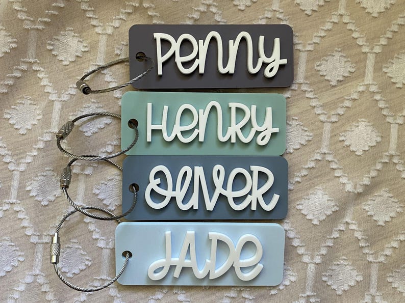 Peut inclure: Quatre &eacute;tiquettes &agrave; bagages color&eacute;es avec des anneaux m&eacute;talliques. Les &eacute;tiquettes sont bleues, vertes, grises et bleu clair. Les noms Penny, Henry, Oliver et Jade sont &eacute;crits en blanc sur les &eacute;tiquettes.