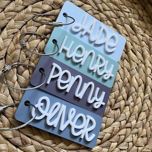 Peut inclure: Quatre &eacute;tiquettes &agrave; bagages de couleur pastel avec des lettres blanches. Les &eacute;tiquettes indiquent "Jade", "Henry", "Penny" et "Oliver". Chaque &eacute;tiquette est munie d'une boucle en m&eacute;tal argent&eacute;.