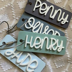Peut inclure: Quatre porte-cl&eacute;s avec des noms diff&eacute;rents. Les noms sont "Penny", "Oliver", "Henry" et "Ade". Les porte-cl&eacute;s sont en acrylique bleu clair et gris avec des lettres blanches. Les porte-cl&eacute;s sont attach&eacute;s &agrave; des anneaux de cl&eacute;s en m&eacute;tal argent&eacute;.