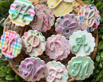 May include: A selection of floral and patterned badge reels, each with a different letter in a cursive font. The reels have scalloped edges and are in various pastel colours, set against a woven basket.