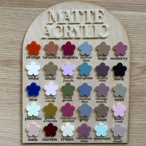 K&ouml;nnte beinhalten: Holz-Displaytafel mit den wei&szlig;en Worten "MATTE ACRYLIC". Die Tafel zeigt ein Raster aus kleinen, bl&uuml;tenf&ouml;rmigen Acrylfarbmustern in verschiedenen Farbt&ouml;nen, darunter Orange, Blau und Rosa. Jede Farbe ist mit ihrem Namen beschriftet.