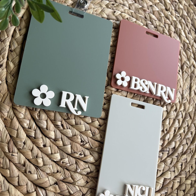 K&ouml;nnte beinhalten: Drei rechteckige Namensschilder in Salbeigr&uuml;n, Staubrosa und Hellgrau. Jedes Schild hat ein wei&szlig;es Blumenmuster und wei&szlig;e Schrift: "RN", "BSNRN" und "NICU". Die Schilder sind auf einem gewebten Hintergrund befestigt.