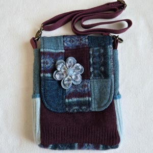 May include: A handmade, patchwork crossbody bag in shades of blue, teal, and burgundy. The bag features a decorative flower and a long, adjustable strap. The bag is made of a soft, textured material.