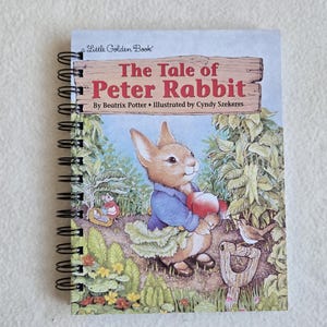 May include: A spiral-bound book titled "The Tale of Peter Rabbit" by Beatrix Potter, illustrated by Cyndy Szekeres. The cover features a watercolor illustration of a rabbit in a blue jacket holding apples, surrounded by greenery.