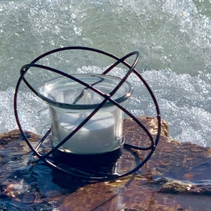 May include: A clear glass candle holder with a white candle inside, encased in a dark metal cage. The candle holder sits on a rock with water and waves in the background. The metal cage has a circular design.