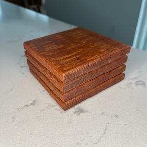 May include: A stack of four square wooden coasters. The coasters are a warm brown colour with a visible wood grain pattern. The edges of the coasters are slightly rounded. The coasters are stacked on top of each other.