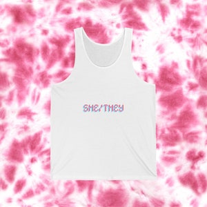 Op de afbeelding: Een wit tanktop met de tekst "SHE/THEY" in een gepixelde, gecrashte lettertype in roze en blauw.