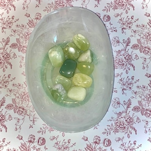 Può includere: Una collezione di pietre di giada verde in una ciotola bianca. Le pietre sono di varie tonalità di verde e hanno una finitura liscia e lucida.