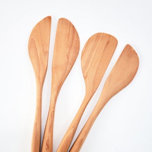 May include: Four wooden spatulas with rounded edges and long handles. The spatulas are arranged in a fan shape, with the handles pointing towards the bottom of the image.