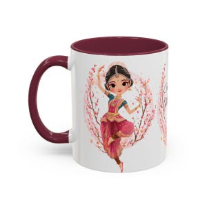 May include: White ceramic mug with a burgundy interior and handle. The mug features a cartoon illustration of a dancing woman in traditional Indian attire, surrounded by pink blossoms. The mug is a standard 11 oz size.