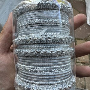 May include: A set of silver bangles with intricate designs and clear rhinestones. The bangles are packaged in clear plastic.