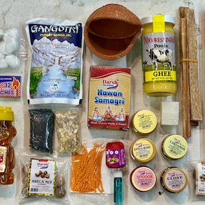 May include: An assortment of items for religious rituals, including ghee, incense, and spices. There's a jar of honey, a clay pot, and a package labeled "Gangotri Purest Ganga Jal." Various small containers and packets are also visible.