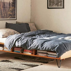 Könnte beinhalten: Ein Bett mit Holzrahmen und eingebautem Bücherregal. Das Bett ist mit einer blauen Bettdecke und Kissen bedeckt. Ein kleiner Teppich liegt vor dem Bett auf dem Boden. Zwei gerahmte Bilder hängen an der Wand.