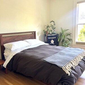 K&ouml;nnte beinhalten: Ein Schlafzimmer mit einem Holzbettgestell und einer dunkelbraunen Bettdecke. Das Bett hat ein wei&szlig;es Kissen und eine wei&szlig;-schwarze Tagesdecke mit Quasten. Im Raum befinden sich Pflanzen und ein kleiner Holz-Beistelltisch.