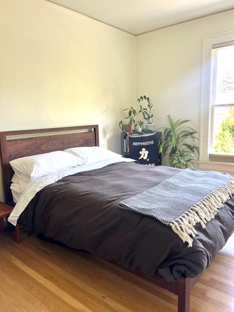 K&ouml;nnte beinhalten: Ein Schlafzimmer mit einem Holzbettgestell und einer dunkelbraunen Bettdecke. Das Bett hat ein wei&szlig;es Kissen und eine wei&szlig;-schwarze Tagesdecke mit Quasten. Im Raum befinden sich Pflanzen und ein kleiner Holz-Beistelltisch.