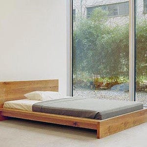 May include: A wooden platform bed frame with a gray mattress and white pillow. The bed is in a room with a large window overlooking a green garden.