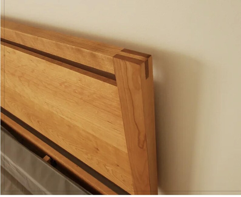 K&ouml;nnte beinhalten: Nahaufnahme der oberen Ecke eines Bettrahmens aus Holz. Der Rahmen besteht aus hellem Holz mit einer dunklen Holzeinlage. Die Ecke ist gefast und mit einer Schwalbenschwanzverbindung verbunden.