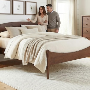 Könnte beinhalten: Ein Schlafzimmer-Set mit einem dunklen Holz-Bettrahmen, einem Nachttisch und einer Kommode. Das Bett ist mit einer cremefarbenen Bettdecke und Kissen ausgestattet, mit einer beigefarbenen Decke. Ein weißer Teppich bedeckt den Boden. Ein Paar steht in der Nähe des Bettes.