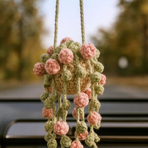 May include: A handmade, hanging crochet ornament featuring a central sphere adorned with pink and green crocheted floral accents. The ornament is suspended by two braided cords, showcasing a blend of textures and colors.