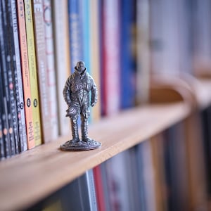 Op de afbeelding: Een zilveren beeldje van een persoon die een jas en broek draagt en op een houten plank staat voor een boekenkast.