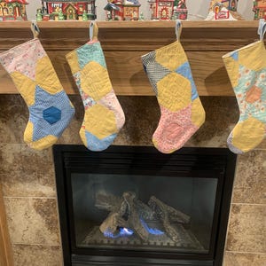 May include: Four patchwork fabric stockings hang on a wooden mantel above a fireplace. The stockings are yellow, blue, pink, and green with a star pattern. The fireplace has a black metal surround and a glass front.