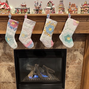 May include: Four white quilted Christmas stockings with floral patterns in shades of blue, pink and green hang on a wooden mantel above a fireplace.