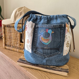 May include: A blue denim tote bag with a large pocket featuring a colorful painted bird design. The bag has a drawstring closure and a brown leather strap.
