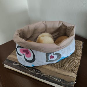 Può includere: Un cestino per il pane in tessuto bianco con una fodera in tessuto marrone e un motivo a forma di cuore. Il cestino è pieno di diversi panini rotondi.