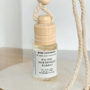 May include: A small, clear glass bottle with a wooden cap and a beige string hanging from it. The bottle contains a clear liquid and has a label that reads "Seven Continents The American Collection Pacific Northwest Forest Oil Diffuser 1 oz (29.5 ml) handmade in the USA".