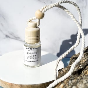 May include: A small glass bottle with a wooden cap and a white rope, labeled "CYPRESS CLIFFS OF BIG SUR." The bottle sits on a white circular platform, with a green sprig and a rock in the background. The background is a marble pattern.