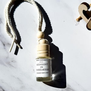 May include: A white string car air freshener with a wooden bead and a clear glass bottle. The bottle has a white label with the text "MIDNIGHT IN MARRAKESH" printed on it.