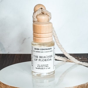 May include: A small, clear glass bottle with a wooden cap and a white label that reads "SEVEN CONTINENTS THE AMERICAN COLLECTION THE BEACHES OF FLORIDA OIL DIFFUSER 0.25 OZ (7ML) handmade in ct us". The bottle is hanging from a white string.