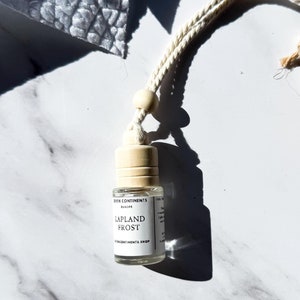 May include: A small glass bottle with a wooden cap and a white label that reads "LAPLAND FROST". The bottle is suspended by a white rope with a wooden bead. A wooden reindeer ornament is visible in the background. The background is a white marble surface.