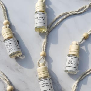 May include: Overhead shot of several small, clear glass bottles with wooden caps and white labels, suspended by cream-colored cords. The labels feature text like "Sangria in Sevilla" and "Italian Aperitivo." The bottles are arranged on a marble-patterned surface.