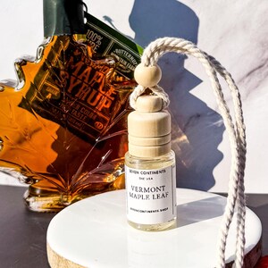 May include: A small glass bottle with a wooden cap and a white label that reads "Vermont Maple Leaf." The bottle hangs from a white cord with a wooden bead. A maple syrup bottle and fall foliage are in the background.
