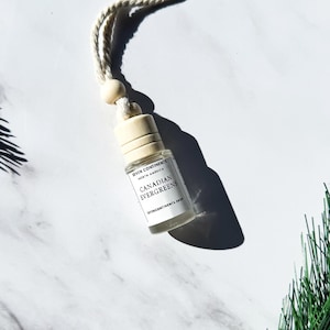 May include: A white car air freshener with a string for hanging. The bottle is labeled "Rhythm & Company" and "Canadian Evergreens" with a black and white design.